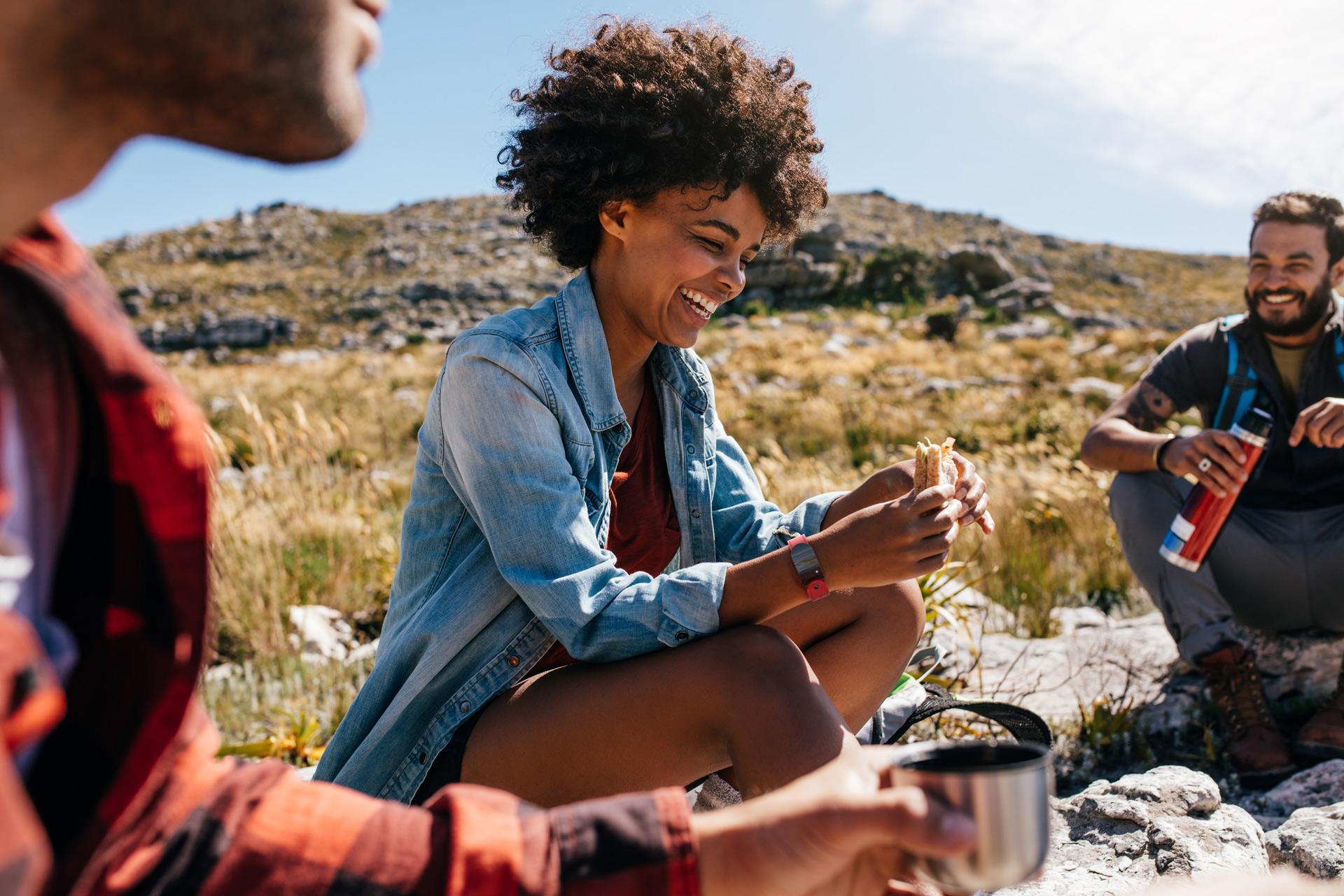Group of happy people during a pic-nic outdoors, a woman in the middle eating a sandwich and wearing a red GEMA Fitness Tracker